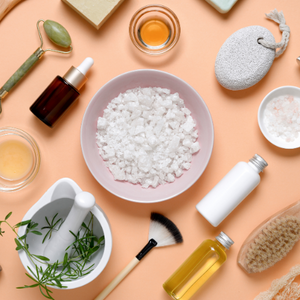 Assorted organic skincare products including sea salt, herbal extracts, oils, and a jade roller on a pastel peach background, representing natural beauty care.